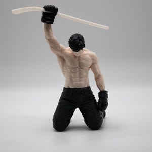May include: A figurine of a muscular person kneeling on the ground with black pants and black gloves. The figure has dark hair and is holding a long, thin, white curved object high above their head with one raised arm in a triumphant pose.