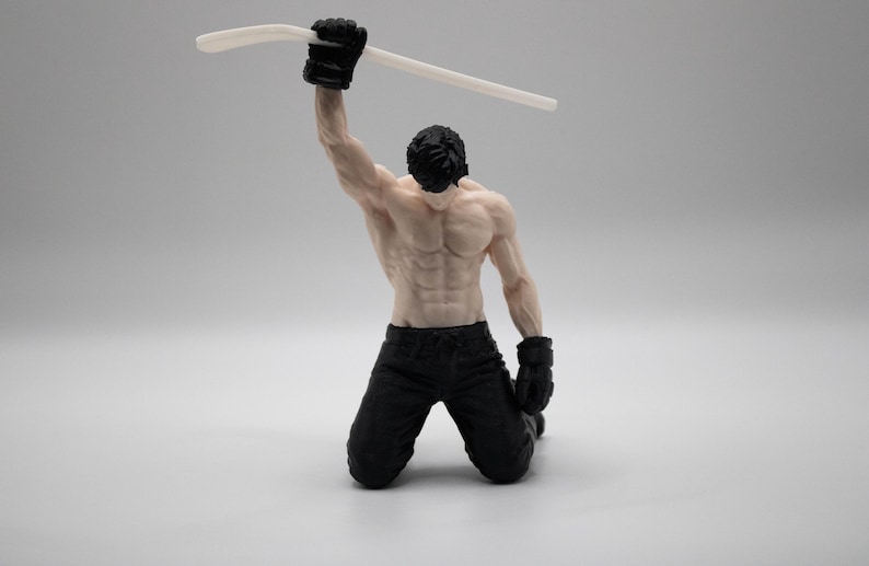 May include: A figurine of a muscular person kneeling on the ground with black pants and black gloves. The figure has dark hair and is holding a long, thin, white curved object high above their head with one raised arm in a triumphant pose.