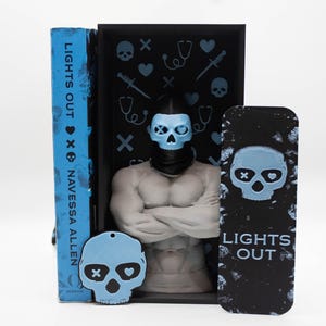May include: A book titled "LIGHTS OUT" by Navessa Allen, with a blue cover, stands next to a black bookend featuring a muscular figure with a skull mask. A matching skull-shaped keychain and bookmark are also present, all with a blue and black color scheme.