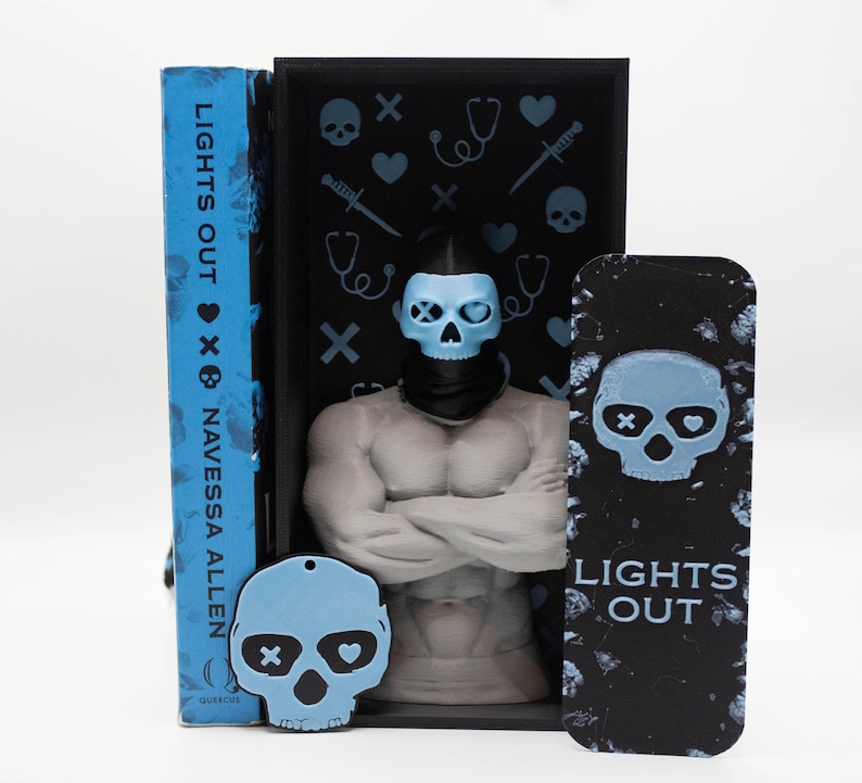 May include: A book titled "LIGHTS OUT" by Navessa Allen, with a blue cover, stands next to a black bookend featuring a muscular figure with a skull mask. A matching skull-shaped keychain and bookmark are also present, all with a blue and black color scheme.
