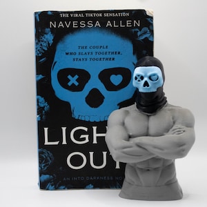 May include: A gray muscular torso figurine with a black balaclava and a light blue skull mask, positioned in front of the book Lights Out by Navessa Allen, which features a large blue skull graphic with an X and a heart over the eye sockets.
