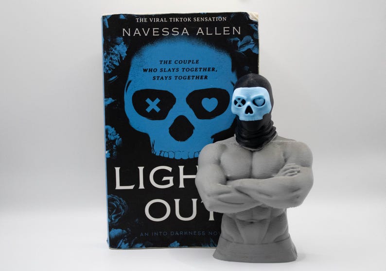 May include: A gray muscular torso figurine with a black balaclava and a light blue skull mask, positioned in front of the book Lights Out by Navessa Allen, which features a large blue skull graphic with an X and a heart over the eye sockets.