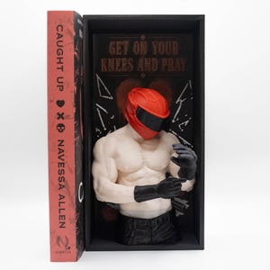 May include: A muscular torso figurine wearing a red motorcycle helmet and black gloves, displayed inside a black box next to a book titled Caught Up by Navessa Allen. The back panel of the box features the text Get On Your Knees And Pray.