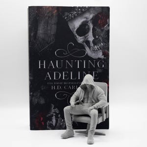 May include: A book titled Haunting Adeline by H.D. Carlton with a black cover featuring a skull and red roses, positioned behind a small, light gray figurine of a person wearing a hooded sweatshirt and pants, seated in a matching chair.