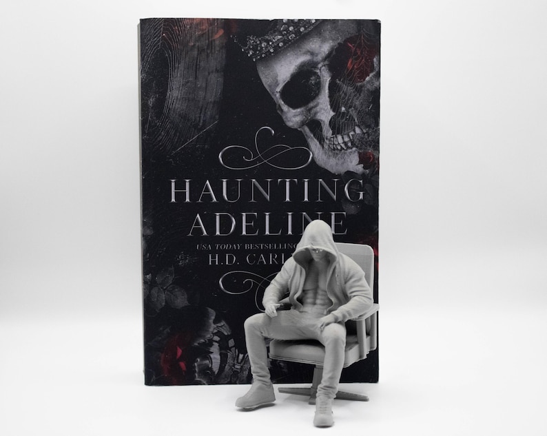 May include: A book titled Haunting Adeline by H.D. Carlton with a black cover featuring a skull and red roses, positioned behind a small, light gray figurine of a person wearing a hooded sweatshirt and pants, seated in a matching chair.