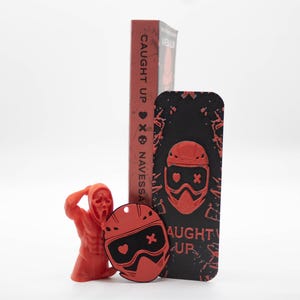 Pode incluir: Uma estatueta vermelha de uma pessoa com uma m&aacute;scara de Scream, um chaveiro vermelho e preto em forma de capacete, uma capa de telefone preta com um design de capacete vermelho e um livro vermelho com o t&iacute;tulo "CAUGHT UP" e o nome do autor "NAVESSA".