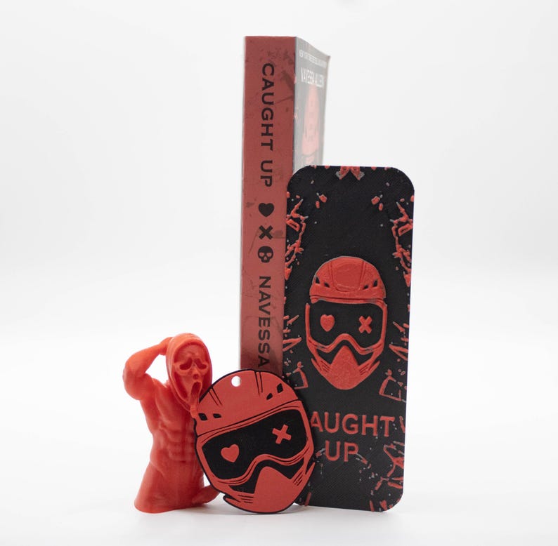 Pode incluir: Uma estatueta vermelha de uma pessoa com uma m&aacute;scara de Scream, um chaveiro vermelho e preto em forma de capacete, uma capa de telefone preta com um design de capacete vermelho e um livro vermelho com o t&iacute;tulo "CAUGHT UP" e o nome do autor "NAVESSA".