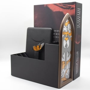 May include: A black display box with a butterfly decoration and a stained glass design. A book with the title "Highest Bidder" is visible. The display box has the Amazon logo.