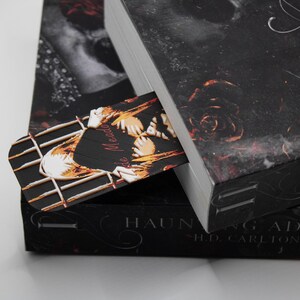 May include: A bookmark with a decorative design is placed inside a book. The bookmark features a black background with a gold and white design. The book has a black cover with a floral pattern and the title "Haunting Adeline".