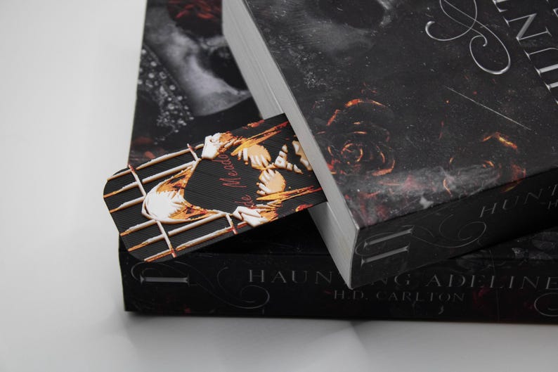 May include: A bookmark with a decorative design is placed inside a book. The bookmark features a black background with a gold and white design. The book has a black cover with a floral pattern and the title "Haunting Adeline".