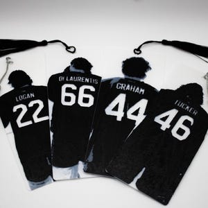 Op de afbeelding: Vier rechthoekige bladwijzers met zwarte sportshirts met witte nummers en namen. De shirts tonen de namen Logan 22, Di Laurentis 66, Graham 44 en Tucker 46. Elke bladwijzer is voorzien van een zwarte of zilveren kwastje aan de bovenkant.