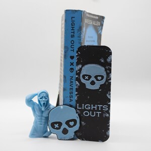 May include: A blue book titled "LIGHTS OUT" by Navessa Allen stands next to a black bookmark with a skull design and the same title. A light blue figurine of a screaming character and a light blue skull-shaped charm are also present.