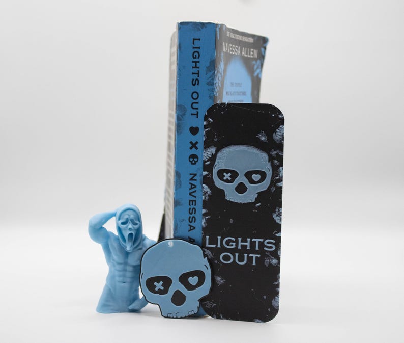 May include: A blue book titled "LIGHTS OUT" by Navessa Allen stands next to a black bookmark with a skull design and the same title. A light blue figurine of a screaming character and a light blue skull-shaped charm are also present.