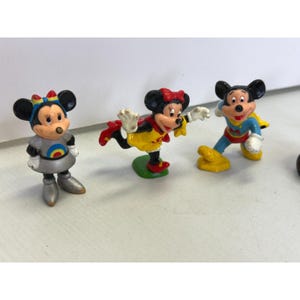 K&ouml;nnte beinhalten: Drei kleine Figuren von Mickey und Minnie Mouse. Eine Minnie Mouse-Figur tr&auml;gt ein silbernes Outfit mit Regenbogenmuster. Eine weitere Minnie Mouse-Figur tr&auml;gt ein gelbes Kleid und rote Schuhe. Eine Mickey Mouse-Figur tr&auml;gt ein blaues und gelbes Superhelden-Outfit.