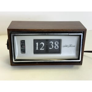 Peut inclure: Une horloge à bascule rectangulaire Seth Thomas avec un boîtier en bois brun foncé et un cadran argenté. L'affichage indique l'heure 12:38 avec des chiffres blancs sur des tuiles noires. Un cadran vertical à gauche indique les chiffres 6 et 7.