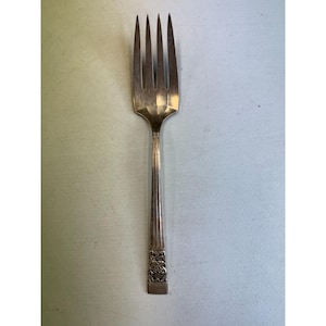 May include: A silver fork with four prongs. The handle features a decorative floral pattern near the base and vertical lines. The fork is made of a shiny, metallic material and is set against a light background.