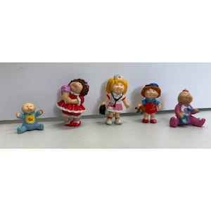 May include: A collection of five small Cabbage Patch Kids figurines. The figurines depict various characters, including a baby in a blue outfit, a girl holding ice cream, a nurse, a boy with a teddy bear, and a baby in a pink outfit.