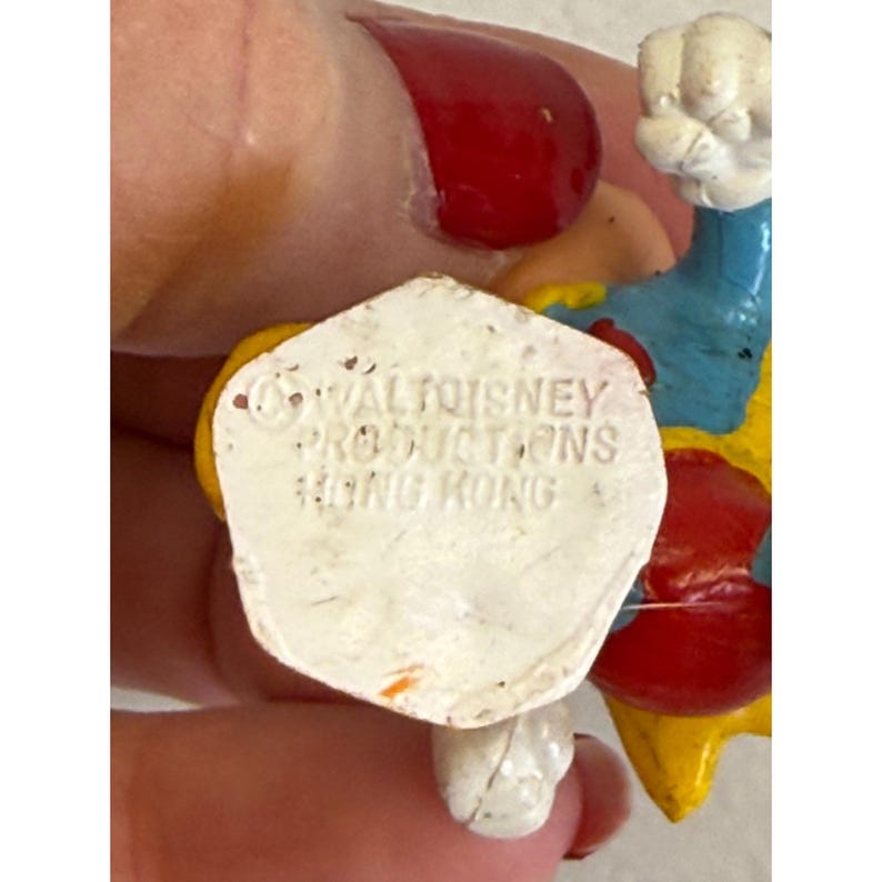 K&ouml;nnte beinhalten: Vintage Figur eines Zeichentrickcharakters in Gelb, Blau, Rot und Wei&szlig;. Der wei&szlig;e Sockel der Figur tr&auml;gt den Text "WALT DISNEY PRODUCTIONS HONG KONG".