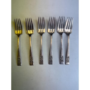 May include: Six silver forks arranged in a row. The forks have four tines and ornate floral designs on the handles. The tines and handles have a shiny, reflective surface. The forks are on a light blue surface.