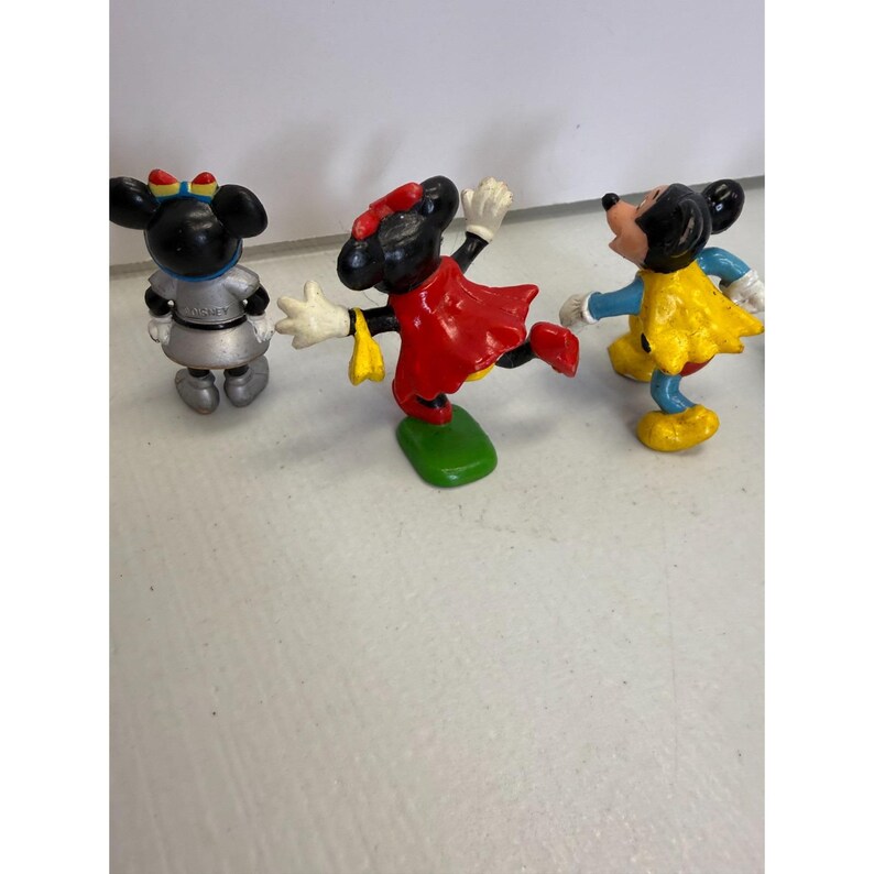 K&ouml;nnte beinhalten: Drei Vintage-Minnie-Mouse-Figuren. Eine ist silberfarben mit einer blauen Schleife, eine andere in einem roten Kleid und die dritte in einem gelben Kleid. Alle haben schwarze Ohren und sind in einer spielerischen Pose. Ein klassisches Disney-Sammlerst&uuml;ck.
