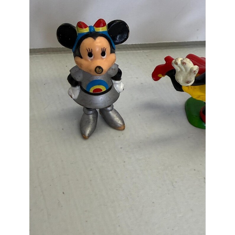 K&ouml;nnte beinhalten: Eine Vintage-Minnie-Mouse-Figur in einem silbernen Raumanzug mit einem blau-gelben Zielscheiben-Design auf der Brust. Die Figur hat schwarze Ohren, eine rote Schleife und steht aufrecht. Die Figur ist ein Sammlerst&uuml;ck.