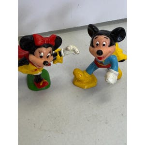 K&ouml;nnte beinhalten: Zwei kleine Figuren von Zeichentrickfiguren. Eine ist eine weibliche Maus mit roter Schleife, gelber Jacke und roten Schuhen. Die andere ist eine m&auml;nnliche Maus in blauer und gelber Kleidung. Beide sind in einer spielerischen Pose.