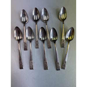 Pode incluir: Uma coleção de nove colheres de prata. Cada colher tem uma tigela oval e um cabo decorativo com um design ornamentado. As colheres estão dispostas sobre uma superfície cinza claro, refletindo a luz.