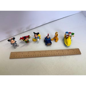 K&ouml;nnte beinhalten: Eine Sammlung kleiner, farbenfroher Disney-Charakterfiguren. Das Set enth&auml;lt Micky und Minnie Maus, Donald und Daisy Duck, Pluto und Goofy. Ein Holzlineal liegt neben den Figuren.