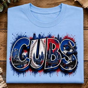 Puede incluir: Una camiseta azul claro con un estampado gráfico de la palabra CUBS en letras grandes. Las letras contienen una textura de pelota de béisbol, un horizonte urbano y una mascota de oso con un bate, todo acentuado por salpicaduras de pintura.
