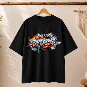 May include: Black t-shirt with a colorful graffiti-style design. The design features the word "SPURS" in bold letters, along with basketball imagery and city skyline elements. The graphic incorporates vibrant colors like orange, blue, and pink.