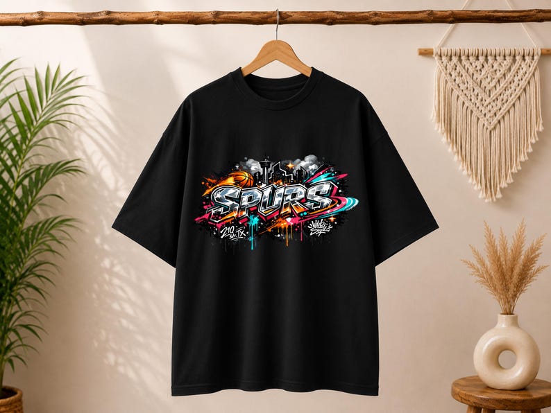 May include: Black t-shirt with a colorful graffiti-style design. The design features the word "SPURS" in bold letters, along with basketball imagery and city skyline elements. The graphic incorporates vibrant colors like orange, blue, and pink.