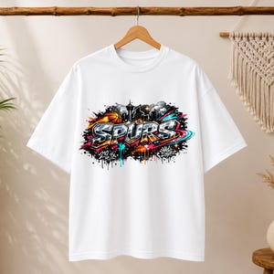May include: White t-shirt with a colorful graphic design featuring the word "SPURS" in graffiti style. The design includes a basketball, cityscape, and the text "210 TX". The shirt is hanging on a wooden hanger.