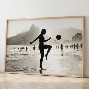 Puede incluir: Impresión enmarcada en blanco y negro de una persona jugando al fútbol en una playa. La silueta de la persona está pateando un balón. Montañas y edificios se encuentran en el fondo, con otras personas en la playa.