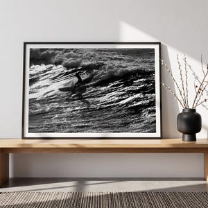 Puede incluir: Una fotografía enmarcada en blanco y negro que muestra a una persona surfeando una gran ola oceánica. La obra de arte se presenta en un marco negro fino, colocada sobre un banco de madera clara junto a un jarrón negro mate con ramas finas.