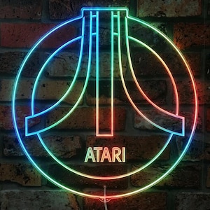 May include: A circular neon sign featuring the Atari logo in a gradient of blue, green, yellow, and red light. The iconic three-pronged symbol is centered within a circular frame, with the word ATARI printed in capital letters below it.