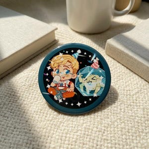 Può includere: Un bottone rotondo color verde acqua con un'illustrazione a cartoni animati. L'illustrazione raffigura un giovane con i capelli biondi, occhiali e una camicia rossa e bianca, che tiene in mano una tazza. Un secondo personaggio è all'interno di un cristallo. Lo sfondo è decorato con stelle bianche e bandiere di festa.