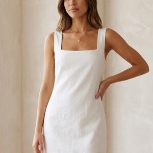 May include: A white sleeveless dress with a square neckline. The dress is knee-length and appears to be made of a linen or cotton fabric. A gold necklace is visible. The model is standing in front of a neutral-colored wall.