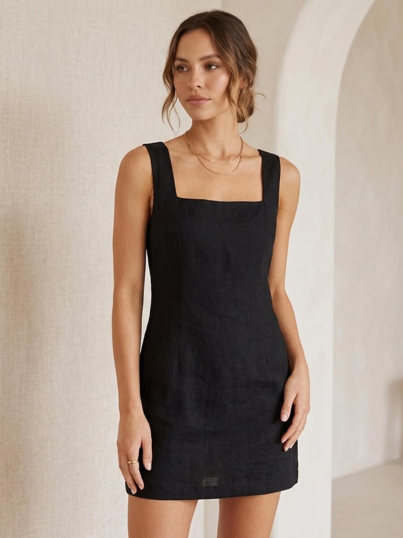 May include: A sleeveless black dress with a square neckline. The dress is knee-length and appears to be made of a textured fabric. A gold necklace is visible around the neck.