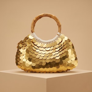May include: A handbag covered in overlapping circular gold sequins that create a reflective, metallic surface. The bag features a round, natural-colored bamboo handle attached to the body with white cord wrapping at the base of the handle.