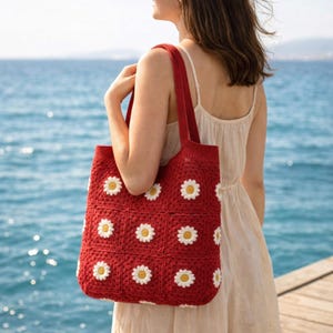 Peut inclure: Un sac fourre-tout rouge en crochet orné de motifs de marguerites blanches et jaunes. Le sac est porté à l'épaule, mettant en valeur sa conception artisanale. L'arrière-plan présente une vue sur la mer.