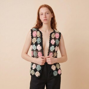 May include: A black crochet vest adorned with colorful floral appliques in shades of pink, green, and beige. The open-front vest is sleeveless and has a scalloped edge. The vest is worn over a black top and black pants.