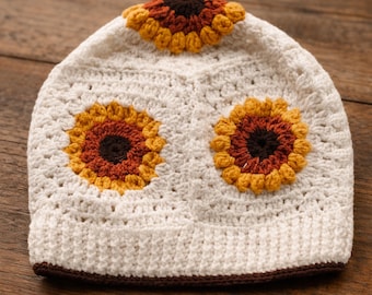 Puede incluir: Un gorro de ganchillo color crema con tres aplicaciones de girasoles. Los girasoles tienen pétalos amarillos, naranjas y marrones con centros marrones. El gorro tiene un borde marrón y se muestra sobre una superficie de madera.