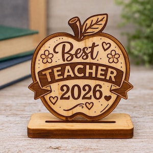 Könnte beinhalten: Ein holzgeschnitztes Apfel-Schreibtischornament mit der Aufschrift "Best Teacher 2026". Der Apfel hat ein Stiel- und Blattdesign mit Herz- und Blumenakzenten. Es steht auf einem kleinen Holzsockel.