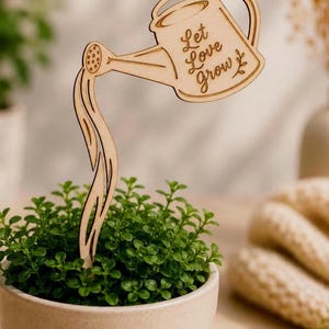 Puede incluir: Una estaca de madera con forma de regadera con la inscripción "Let Love Grow". Está insertada en una maceta pequeña, redonda y beige llena de plantas verdes. La regadera está vertiendo un chorro de agua.