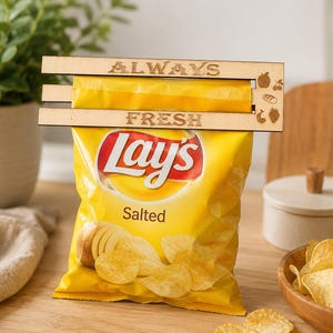 Puede incluir: Un clip de madera de color claro con el texto grabado a láser ALWAYS FRESH y pequeños iconos de una manzana, una cereza, pan y una fresa. El clip está sujeto a la parte superior de una bolsa amarilla de patatas fritas Lay's Salted.