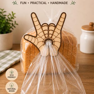 May include: A wooden bread bag clip in the shape of a hand with a spiderweb design, holding a clear plastic bag of sliced bread. The text "BREAD BAG CLIP" is at the top, with "FUN • PRACTICAL • HANDMADE" below it. The clip is eco-friendly and made of natural wood.