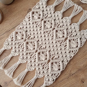 Pode incluir: Um caminho de mesa de macramé em tom off-white com um padrão de diamante repetitivo criado com cordão intrincadamente atado. A peça retangular é finalizada com longas franjas retas nas bordas superior e inferior, exibindo um design texturizado.