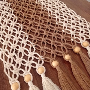 Puede incluir: Camino de mesa de macramé con un diseño geométrico. El camino de mesa es bicolor, con secciones en blanco roto y marrón. Cuentas de madera y borlas adornan los extremos. Adecuado para la decoración del hogar.