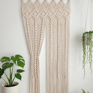 Op de afbeelding: Een crèmekleurige macramé wandhanger met een ingewikkeld geometrisch ruitpatroon aan de bovenzijde, overgaand in lange, verticale franjes met kleine knoopaccenten, opgehangen aan een horizontale houten roede.