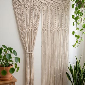 May include: Cream macrame curtain featuring diamond and beaded designs, hung from a wooden rod. The curtain is tied back on the left. Potted plants are seen on either side of the curtain, adding a natural touch.